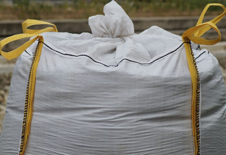 Stor vit byggsäck med gula lyftöglor fylld med byggavfall på en utomhusarbetsplats.